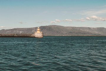 Yellow lighthouse in ocean bay against blue sky and mountains on horizon. Copy space.