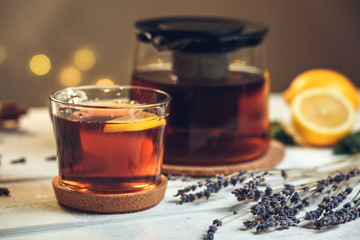 One full Cup of black tea with lemon and teapot, with lavender, dry cloves, fresh mint and cinnamon sticks, on a white wooden table on a background of bokeh lights, still life