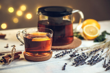 One full Cup of black tea with lemon and teapot, with lavender, dry cloves, fresh mint and cinnamon sticks, on a white wooden table on a background of bokeh lights, still life
