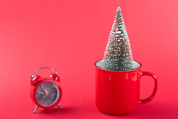minimal christmas concept, a christmas tree sticks out of a red cup, next to a red alarm clock, on a red background, copy space
