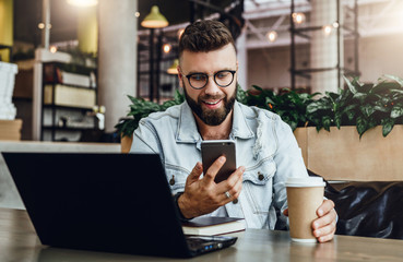 Bearded businessman sitting in cafe, talking on smart phone, working on laptop, freelancer working...