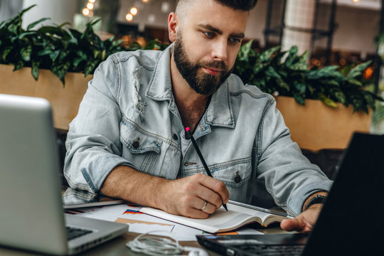 Serious bearded hipster man sitting in office, making notes in notebook, working. Entrepreneur analyzes information.