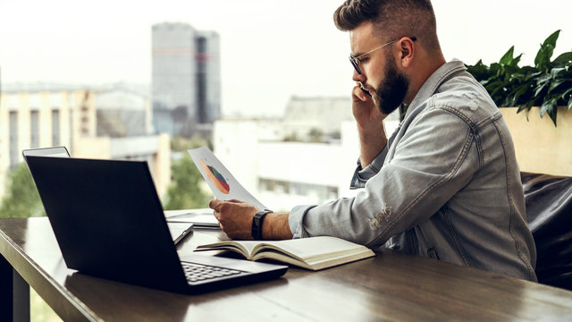 Bearded businessman hipster talking on phone while sitting at desk in office, sad looking at documents. Fall in profits.