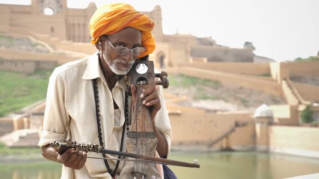 An Old traditional man playing Sarangi (An String instrument) against the historic fort. An elderly street male musician in pagri playing an Indian classical music instrument beside lake in Rajasthan 
