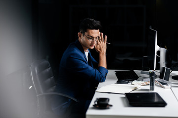 The architect in glasses dressed in a blue jacket sits at the desk in front of the computer and thinking