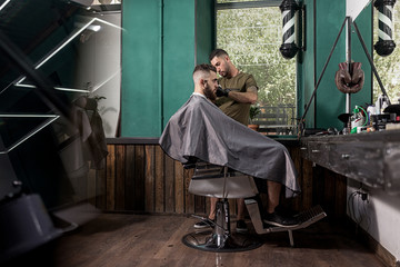 Brutal man with tha beard sits in the chair in front the mirror at a barber shop . Barber shaves...