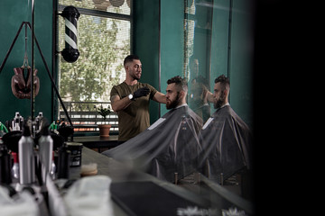 Man with a beard sits in the chair in front the mirror at a barber shop. Barber makes a hairstyle.