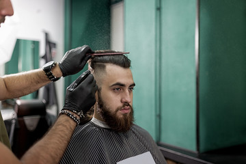 Stylish man with a beard sits at a barber shop. Barber in black gloves does styling and spraying for him