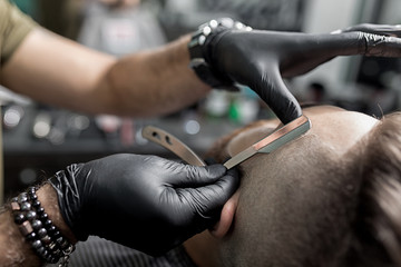 Barber in black gloves trims temple of brutal bearded young man with a straight razor at a...
