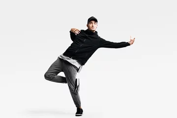 Gordijnen Handsome young man wearing a black sweatshirt, gray pants and a cap dancing street dances on a white background © Leika production
