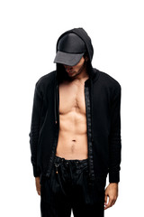 Naklejka na ściany i meble Handsome young dancer dressed in a sweatshirt on a naked torso with a hood on the cap and black pants stands on a white background