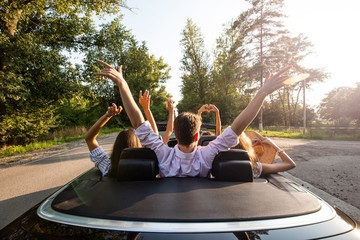 Сompany of young people riding in a cabriolet on the road and holding their hands up on a warm sunny day. Back view.