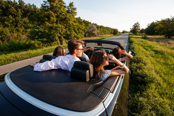 Company of young girls and guys are sitting in a black cabriolet on the country road on a sunny day.