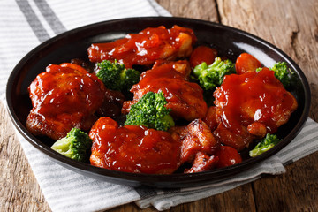 Chicken thighs in red sweet-sour sauce served with broccoli and tomatoes close-up on a black plate. horizontal
