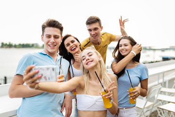 A company of good-looking friends laughing and drinking yellow cocktails and socialising and making selfie in the nice cafe next to the river. Cheers. Entertainment, having good time. River is in the