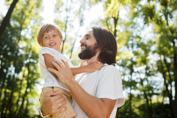 Naklejka premium Handsome dark-haired father with beard dressed in the white t-shirt is holding in the arms his little son and watching at him on a summer day in the park.