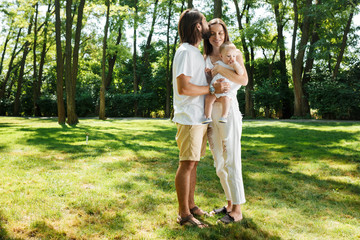 Fototapeta premium Dark-haired father kisses his beautiful wife holding a small daughter in a white dress in a park on a sunny day.