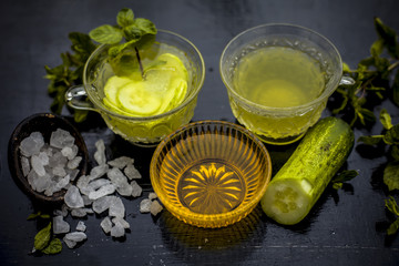 Iced cucumber and mint tea on wooden surface in a transparent cup with slices of cucumber and raw cucumber,mint leaves,sugar and honey with green tea in a separate glass cup.