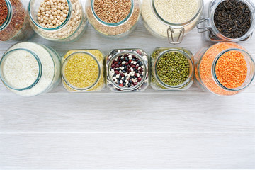 Obraz na płótnie Canvas Various cereals, grains and beans in glass jars. Lentils, chickpeas, peas, beans, quinoa, rice and bulgur. Concept Clean eating. White background and wooden table 