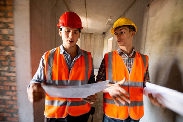 Two men dressed in orange work vests and helmets work with construction documentation inside the...