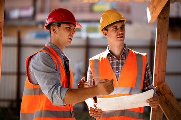 Two men dressed in shirts, orange work vests and helmets explore construction documentation on the...