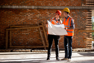 Structural engineer and architect dressed in orange work vests and helmets discuss documentation on...
