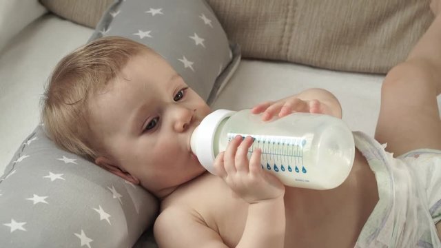 Close up of little 1 year old baby in diaper holds sucking bottle of milk in hands, looks at camera and drinks infant formula 