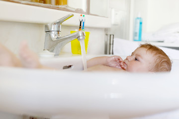 Cute adorable baby taking bath in washing sink and grab water tap.