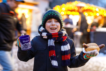 Little cute kid boy eating German sausage and drinking hot children punch on Christmas market
