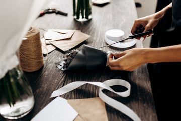 Florist cuts a ribbon on the little bouquet of flowers packed in dark paper on the wooden table with postcard, envelopes and white ribbon on it