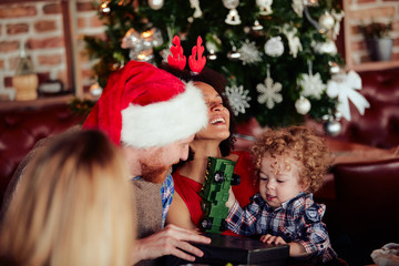 Fototapeta na wymiar Friends giving gift to toddler while sitting at table. Christmas tree in background. Christmas holiday concept.