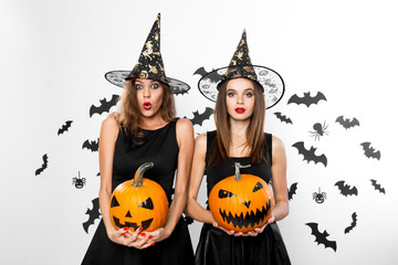 Two brunette girls in black dresses and witch`s hats hold horror halloween pumpkins on the background with bats