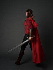 full length portrait of brunette girl wearing red medieval costume and cloak. standing pose  with back to the camera, holding a sword on grey studio background.