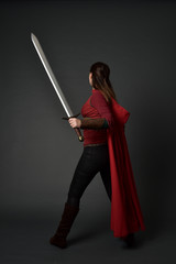 full length portrait of brunette girl wearing red medieval costume and cloak. standing pose  with back to the camera, holding a sword on grey studio background.