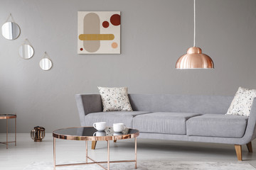 Copper lamp and coffee table in front of a modern sofa in a grey living room interior. Real photo