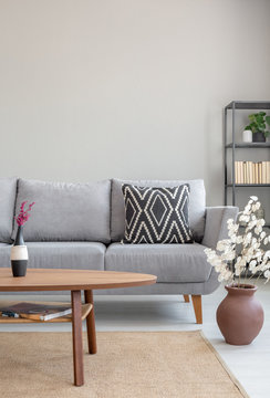 Wooden table in front of grey settee in simple apartment interior with flowers and carpet. Real photo