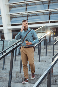 handsome middle aged businessman holding coffee to go and talking by smartphone on stairs