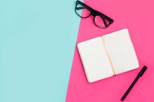 Flat lay, top view office table desk. Work space with open Notepad, glasses and pens on pink and blue paper background