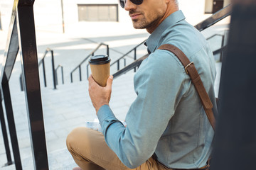 cropped shot of man in sunglasses holding paper cup with coffee to go while sitting on stairs