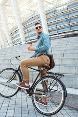 stylish man in sunglasses sitting on bike and holding disposable coffee cup