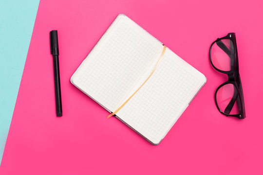 Home office desk with notepads, pen, glasses on a pink background
