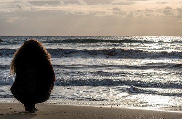 A Beautiful girl sitting on a sandy beach in silence with a backpack. Thinking. Horizontal. Back View