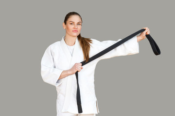 Confident attractive young athletic woman in white kimono standing and showing black belt, looking...