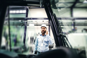 A portrait of an industrial man engineer with smartphone in a factory, making a phone call.