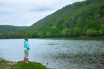 female traveller in hat and rubber boots stands near the river and watching at the hill on another bank