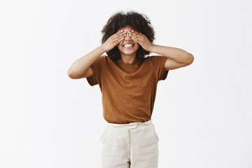 Studio shot of bright happy and young attractive african american short girl with afro hairstyle covering eyes with palms and smiling carefree as if playing hide n seek being in playful mood