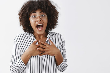 Fototapeta na wymiar Waist-up shot of touched surprised and grateful attractive young female coworker with dark skin, afro hairstyle in striped blouse holding hands on breast and smiling with opened from amazement mouth