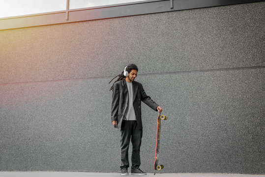 Modern urban young dreadlocks hipster man holding his skate on street and listening music. Standing against black wall with headphones on his head.