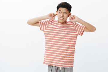 Fototapeta premium Young guy cannot focus on homework as hearing irritating and disturbing loud noise coming from upstairs clenching teeth and frowning annoyed, closing ears with index fingers, looking up angry