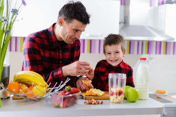 Proud young good looking father and his cute son together in kitchen making smoothie with fresh fruits. Healthy eating is important.